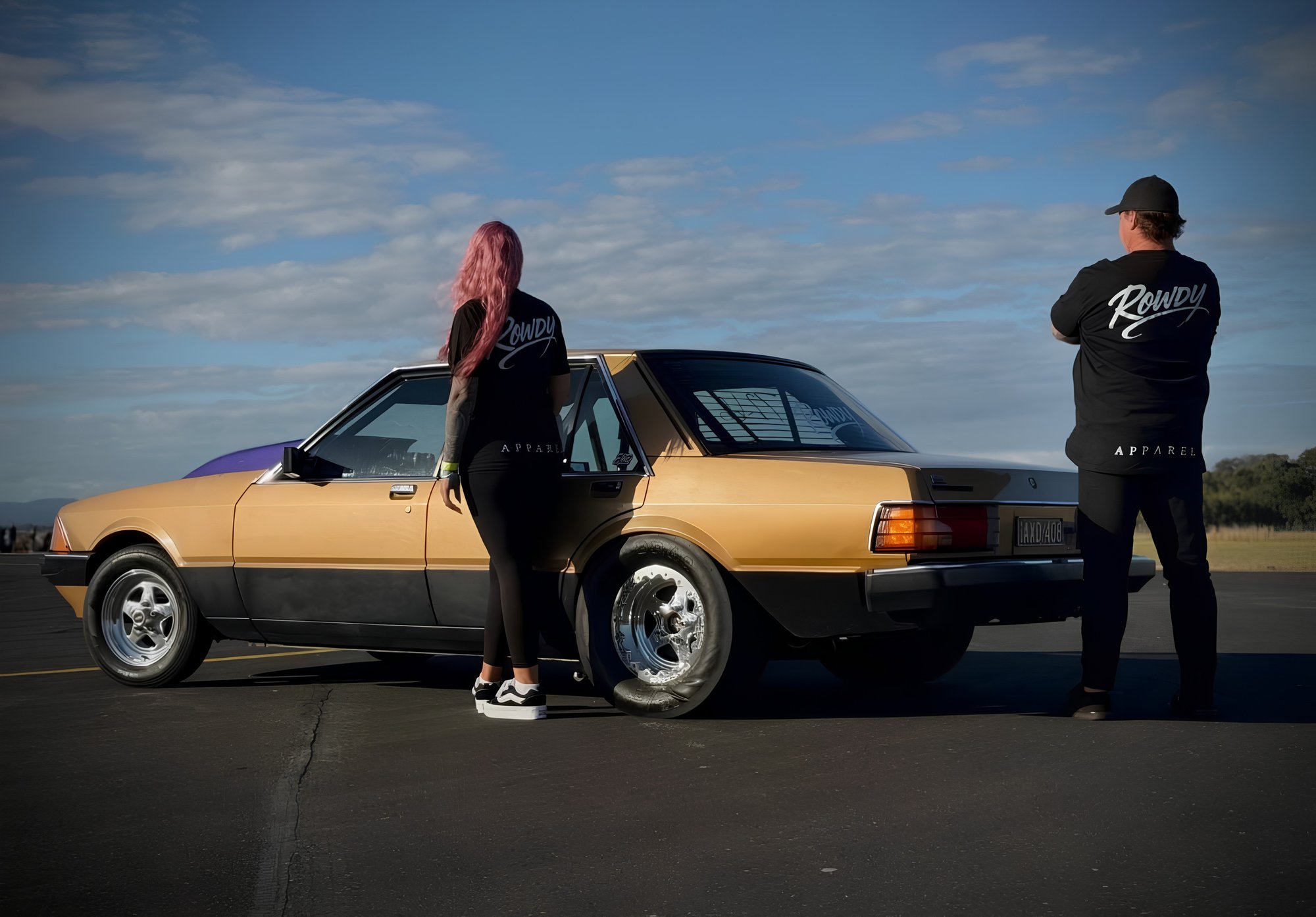 Two people in Rowdy Apparel tees beside a gold Ford Falcon on the tarmac, sun low