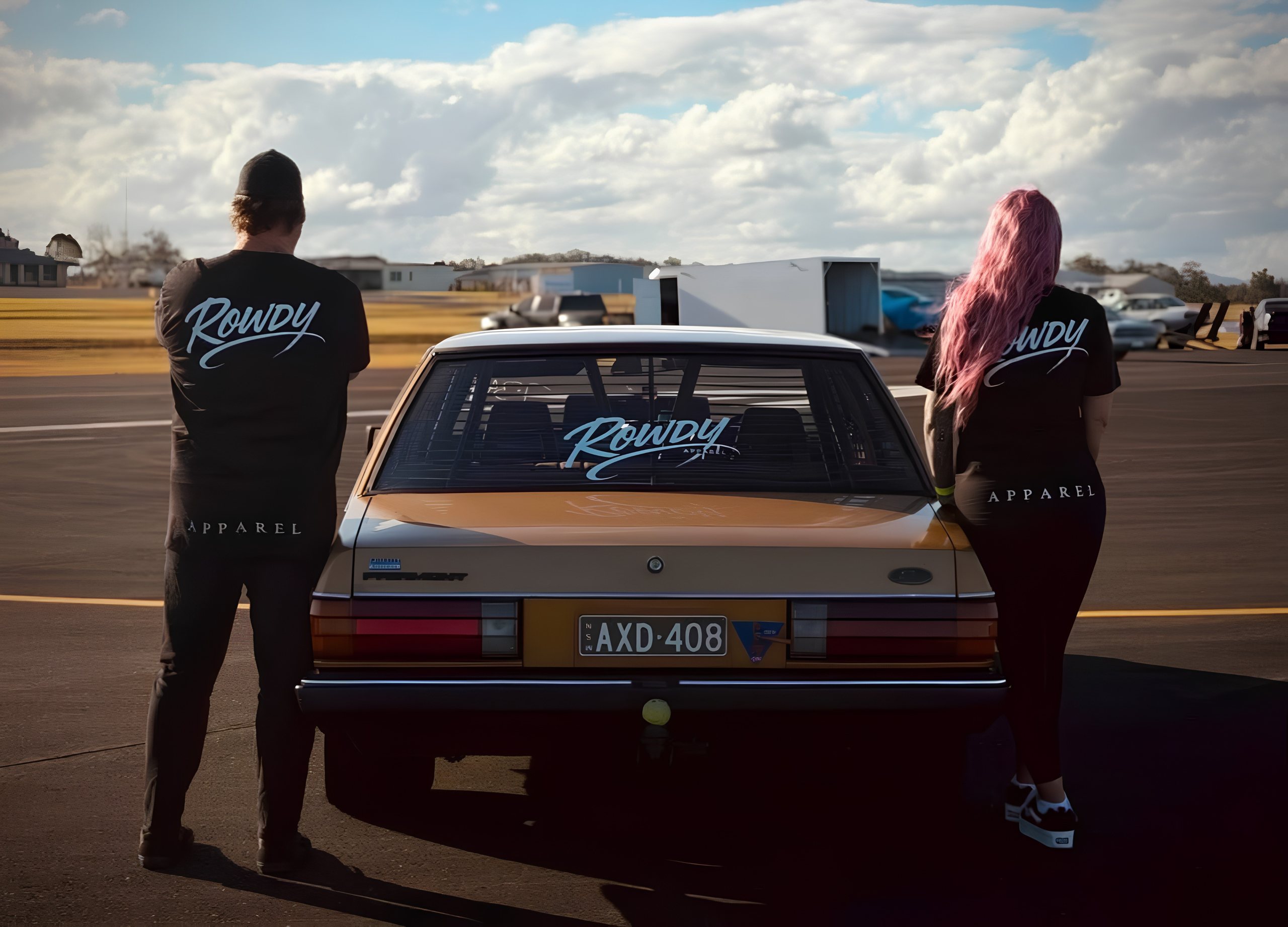 Two Rowdy Apparel crew members in branded tees standing behind a gold Ford Fairmont at a regional airfield drag strip.
