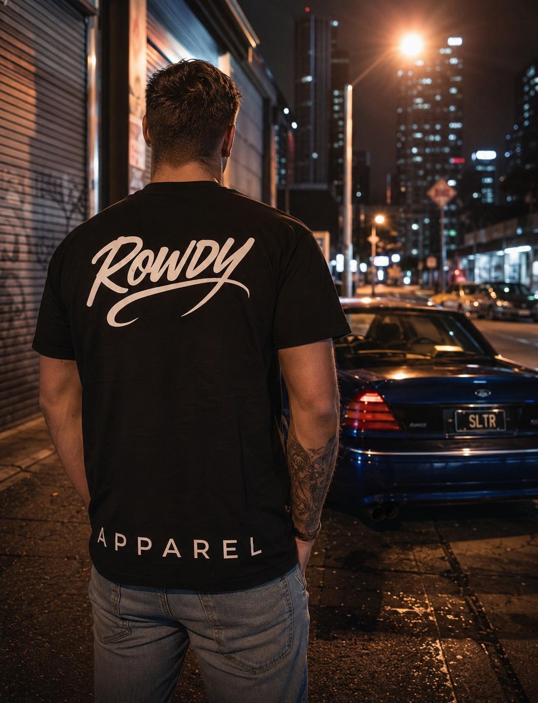 The black Rowdy tee worn back-on at night on a city street, script print across the shoulders