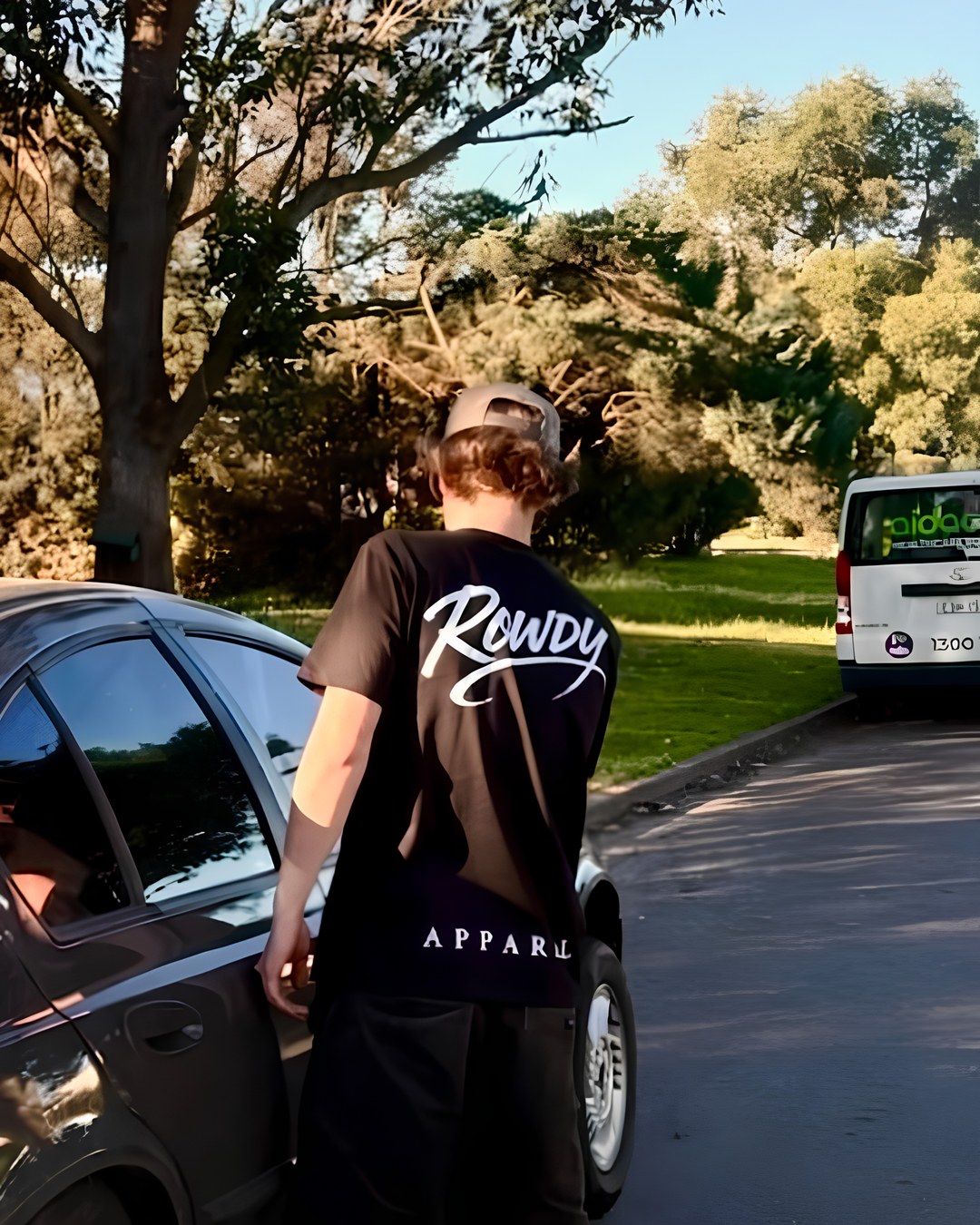 The black Rowdy tee worn back-on beside a car on a tree-lined street, script print across the shoulders and APPAREL along the hem