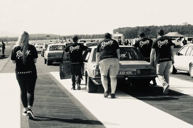 Black and white photo of the Rowdy crew in matching branded tees walking down a drag strip toward a car.